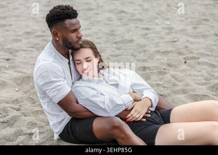 A couple relaxes on the sandy beach, embracing gently. The man sits with his back to the sun, holding the woman close as she leans her head on his sho Stock Photo