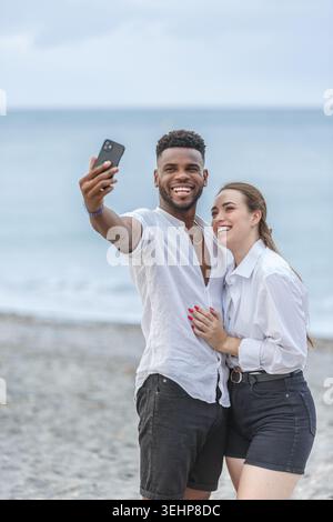 A joyful couple stands close together on the sandy shores of Playa del Zapillo in Almeria, Spain. They are taking a selfie, both smiling widely, with Stock Photo