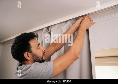 Focused young bearded man hanging curtains on rod during apartment cleaning, organizing home, performing household chores, doing routine house Stock Photo