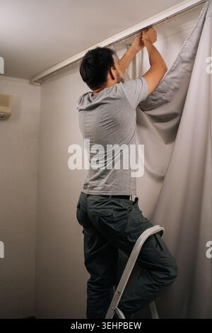 Vertical shot of man standing on a stepladder, busy installing new curtains on rail, performing household chores and home maintenance, focusing on Stock Photo