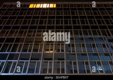 Minimalist night city photo: single warm-lit window glows alone at 6 AM in a vast dark modern glass office facade. Quiet, empty streets. Stock Photo
