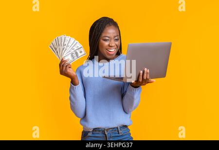 Young Woman Smiles While Holding Money and Looking at Laptop With Excitement Stock Photo