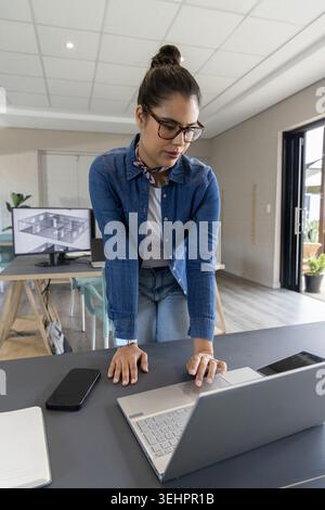 Woman in denim leaning over conference table in office reviewing 3D plans on laptop with smartphone Stock Photo