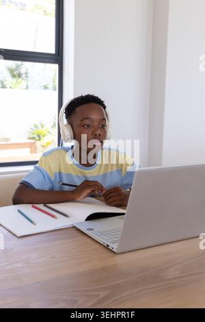 African American boy sitting at table using laptop wearing white headphones, writing in notebook Stock Photo