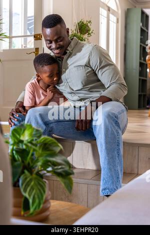 African American father and son sitting on steps holding close at home entryway, glass-paneled door Stock Photo