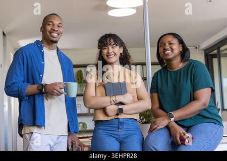 Diverse coworkers posing and smiling at modern office island, holding tablet, phone, mug Stock Photo