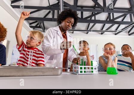African American teacher in lab coat guiding children at lab table using dropper test tubes goggles Stock Photo