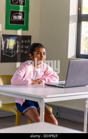 School-age Asian girl sitting on yellow chair in classroom, using silver laptop, wearing pink top Stock Photo