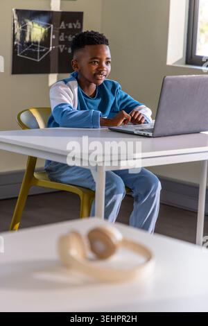 African American preteen boy typing on laptop at classroom table with headphones nearby Stock Photo