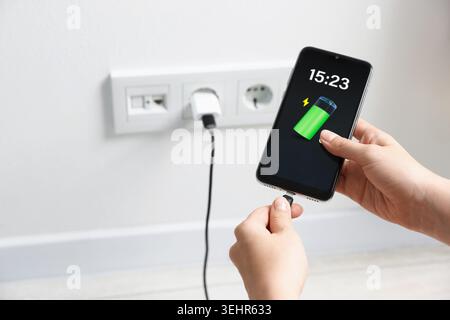 Woman connecting charging cable to smartphone indoors, closeup. Display showing battery level Stock Photo