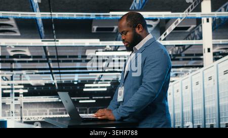 Using laptop, workspace desk and technician in server hub doing maintenance on machines. Configuring server settings, notebook and African american worker in modern high tech data center, camera B Stock Photo