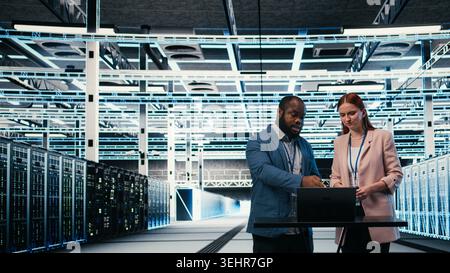 Data center coworkers collaborating to manage enterprise cloud computing IT solutions using laptop. Team of programmers discussing, monitoring futuristic server farm facility workspace, camera A Stock Photo
