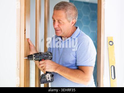 Mature man himself installs the door hinges with screwdriver Stock Photo