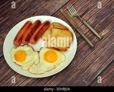 Two fried eggs, fried sausages, and toasted bread on a round white plate on a wooden table, with cutlery nearby. Top view, flat lay. Stock Photo