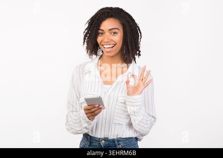 Portrait of smiling afro girl using phone showing ok gesture Stock Photo