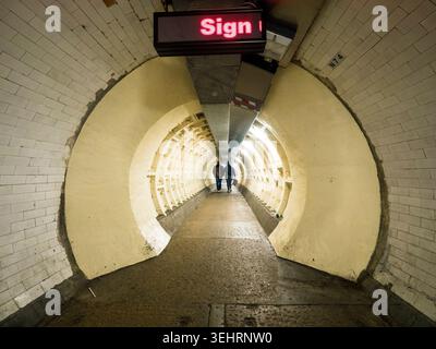 A couple walks hand-in-hand through the historic Greenwich Foot Tunnel beneath the River Thames, curved white-tiled walls receding into the distance Stock Photo