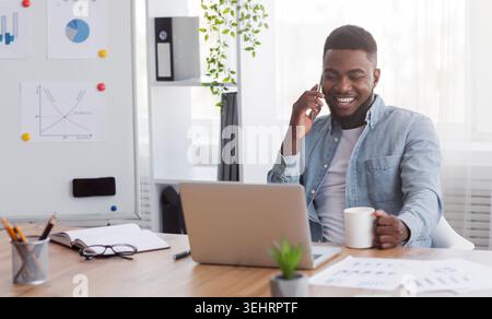 Cheerful black manager talking on mobile phone and drinking coffee Stock Photo