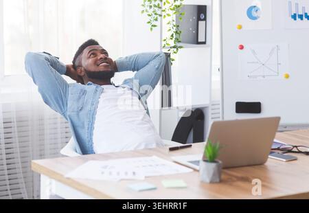 Dreamy black employee resting at workplace with hands behind head Stock Photo