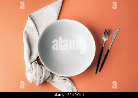 Minimal table setting with empty ceramic plate, linen napkin and modern cutlery on warm terracotta background. Elegant dining composition with copy sp Stock Photo