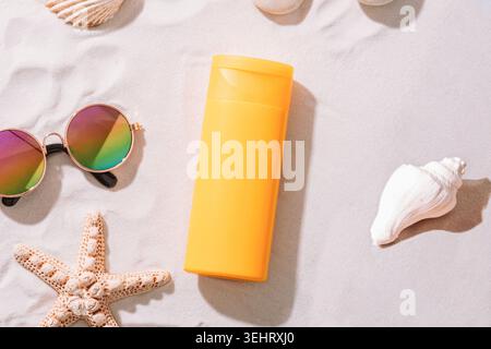 Yellow sunscreen bottle with sunglasses, seashells and starfish on white sand beach background. Summer skincare and sun protection concept. Top view, Stock Photo