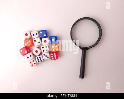 A cluster of colorful gaming dice sits next to a black magnifying glass on a plain white background. Stock Photo