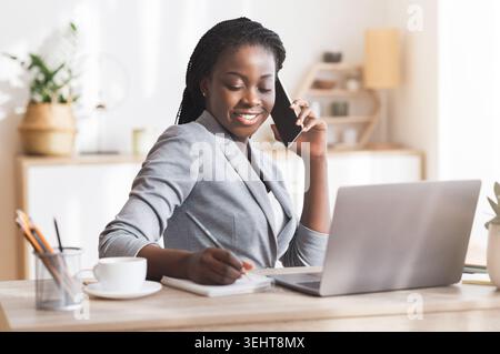 Afro Businesswoman Taking Notes While Having Phone Conversation In Modern Office Stock Photo