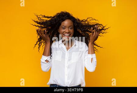 Smiling Woman With Curly Hair Enjoying Vibrant Yellow Background While Playing With Her Hair Stock Photo
