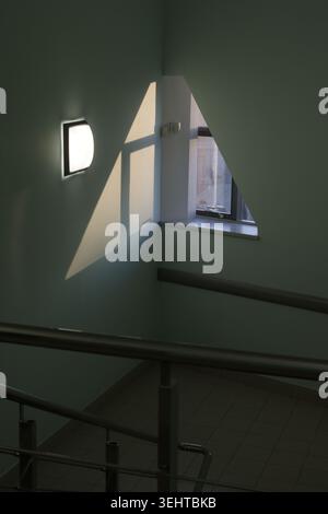 Metal railings frame a quiet staircase. Walls glow pale under filtered daylight. Shadow lines dance across smooth surfaces Stock Photo
