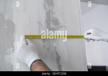 Measuring tape in gloved hands is positioned against a textured wall surface during inspection at a construction site. Clean environment with visible texture and color variations on the wall. Stock Photo