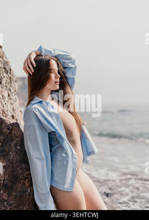 Woman beach shore beautiful young woman with long hair wearing open light blue shirt standing by rock on coast Stock Photo