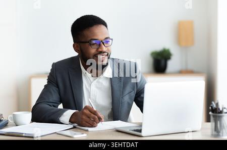Handsome black entrepreneur working on laptop and taking notes Stock Photo