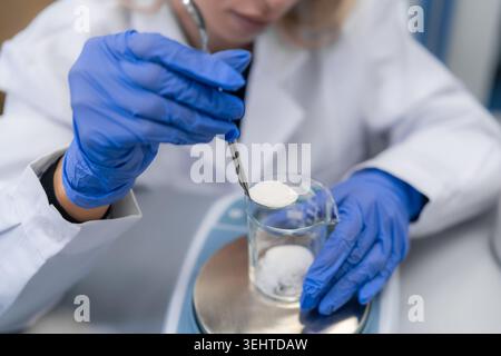 Conducting precise measurement of a powder sample using a spoon over glass beaker in pharmaceutical research lab. The technician wears blue gloves and a lab coat, ensuring safety and accuracy. Stock Photo
