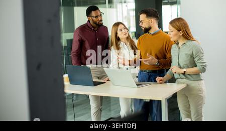 Four people are gathered around a table in an office, talking and sharing ideas about a project. Two laptops are open and a cup of coffee is nearby on Stock Photo