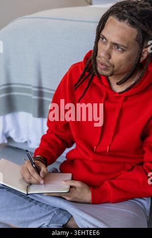 African American man sitting on bed, writing in notebook with black pen, wearing red hoodie Stock Photo