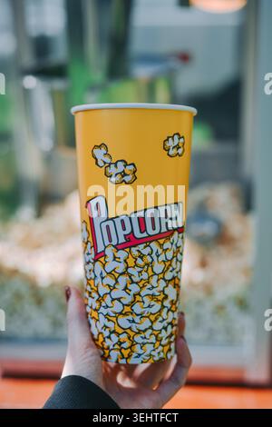 Riga, Latvia -May 23, 2026: Close-up of a hand holding a colorful popcorn cup with blurred snack stand background. Bright daylight, shallow depth of f Stock Photo