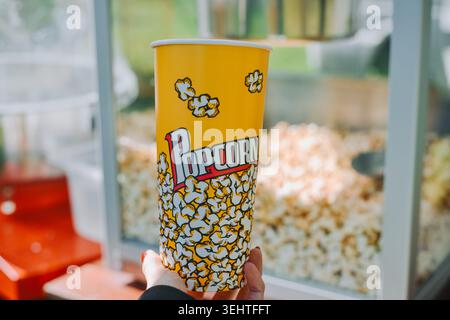 Riga, Latvia -May 23, 2026: Close-up of a person holding a yellow popcorn cup in front of a popcorn machine at a movie theater concession stand. Brigh Stock Photo