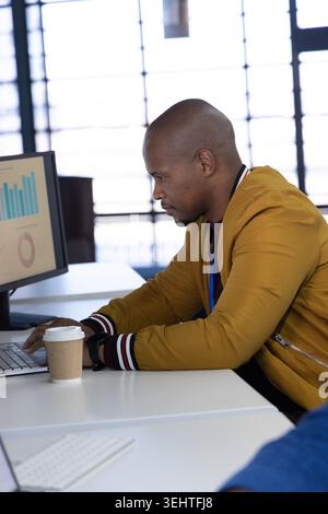 African American man wearing mustard jacket typing on laptop at desk with charts and coffee cup Stock Photo