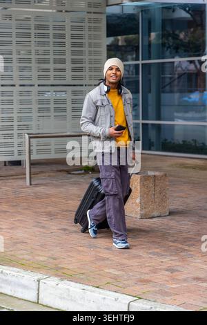 Man walking across brick plaza pulling black suitcase and holding smartphone wearing denim jacket Stock Photo