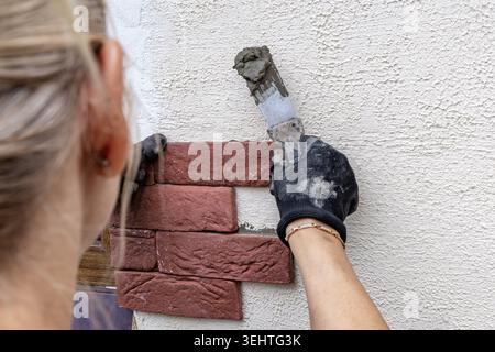 Renovation of the exterior facade of a summer house in the garden, finishing work on laying tiles and gluing them to the facade, a woman gluing red br Stock Photo