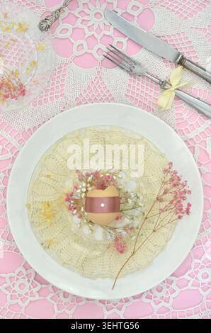 Vintage, feminine Easter table setting: plate, crocheted napkins, Easter egg, retro fork and knife, bow, pink flowers and other romantic decorations. Stock Photo
