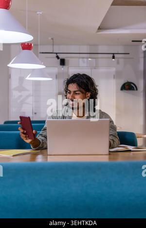 Indian man mid 30s smiling, checking red smartphone and typing on silver laptop in coworking lounge Stock Photo