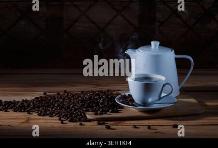 Moody still life of steaming hot coffee served in a white ceramic cup and teapot with scattered roasted coffee beans on a rustic wooden board. Stock Photo