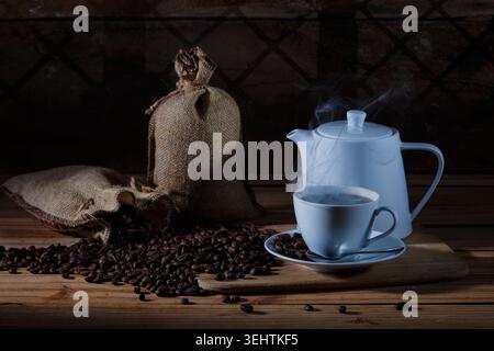 Moody still life of freshly brewed hot coffee in a white ceramic cup and teapot with steaming vapor, surrounded by scattered roasted coffee beans spil Stock Photo