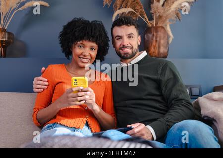 Happy multiracial couple sitting on sofa and looking at smartphone together at home Stock Photo