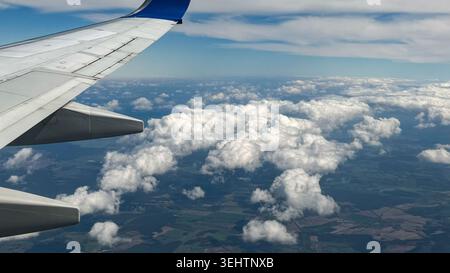 Airplane wing extending over fluffy white clouds with a clear blue sky and green fields visible below, capturing a serene aerial view from a flight Stock Photo
