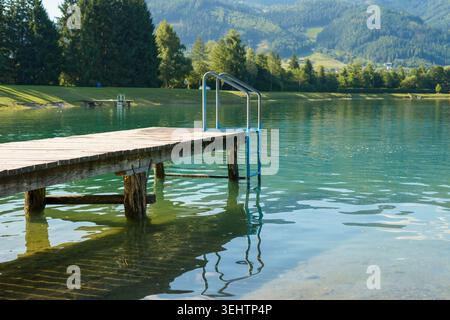 Peaceful lake scene with wooden pier and metal ladder, surrounded by green forest and hills, perfect place for relaxation, swimming and nature escape Stock Photo