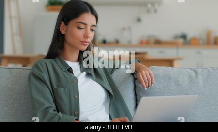 Pensive Indian woman using laptop computer thinking typing freelancer at home student Arabian female ethnic girl businesswoman browsing online Stock Photo