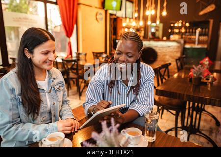 Diverse friends connecting and smiling using digital tablet in cafe Stock Photo