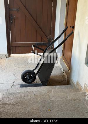A black metal wheel barrow positioned leaning against a white wall on a stone covered pathway in a Bulgarian house Stock Photo