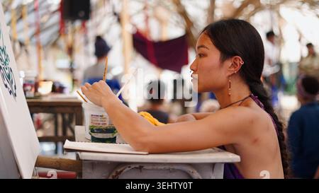 Young woman creating art on a canvas, holding a paintbrush at an outdoor event Stock Photo